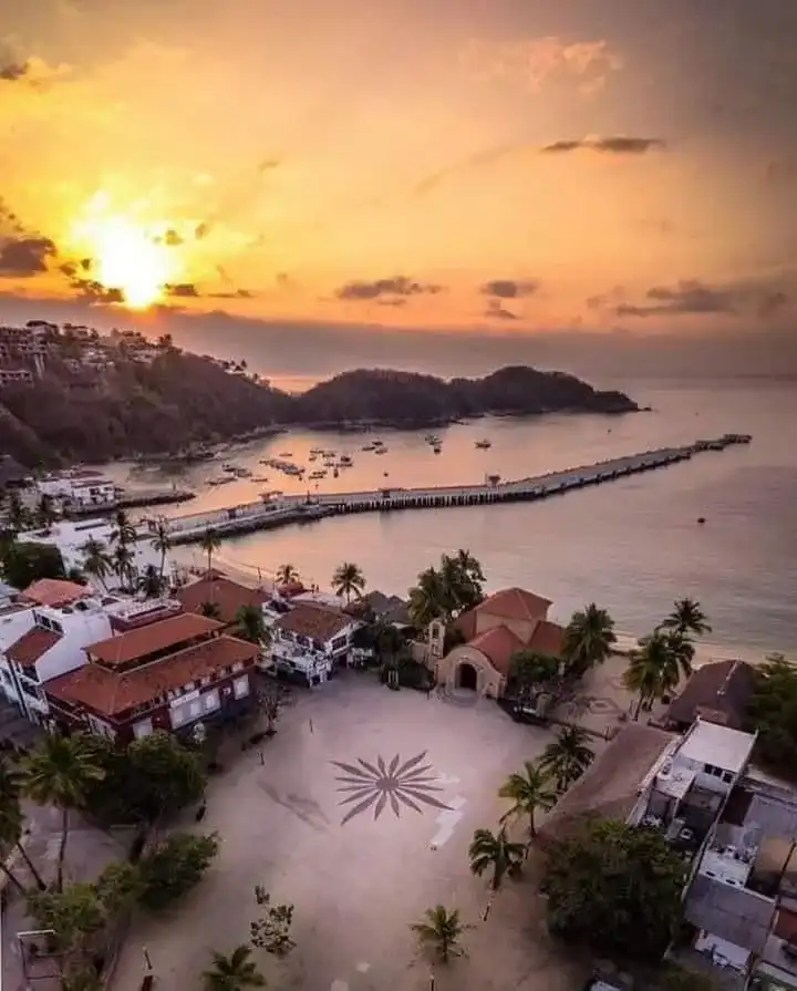 Atardecer naranja sobre una bahía de Huatulco vista desde lo alto, con un muelle y palmeras