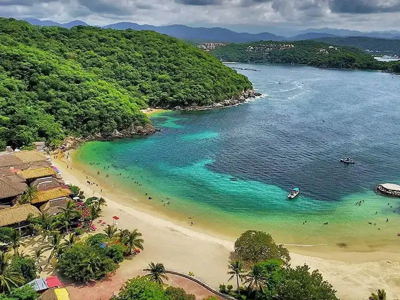 Vista aérea de una bahía de Huatulco con agua turquesa, arena clara y selva en los cerros