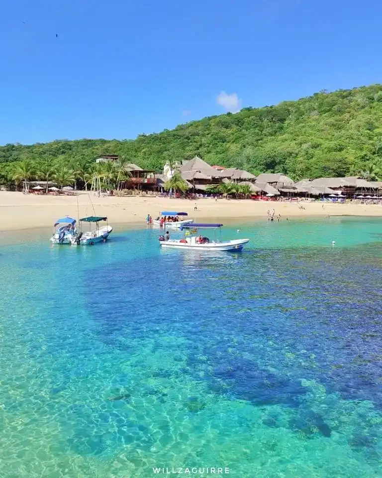 Lanchas fondeadas en una bahía de agua turquesa frente a una playa con palapas